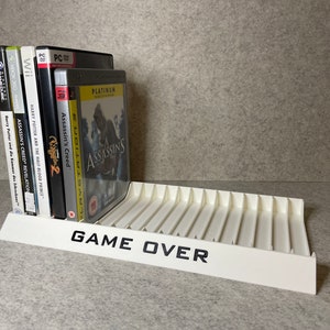 Puede incluir: Estante de almacenamiento de juegos de video de plástico blanco con el texto "GAME OVER" impreso en la parte delantera. El estante contiene una variedad de cajas de juegos de video, incluyendo Assassin's Creed, Harry Potter y otros títulos.