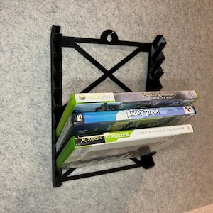May include: Black wall-mounted video game storage rack with four video game cases. The cases are for the games "Pirates of the Caribbean: At World's End", "Angry Birds", "The Elder Scrolls III: Morrowind", and "Xbox Classics".