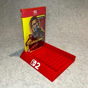 Könnte beinhalten: Ein roter Switch-Spielkofferhalter mit einem Spielkoffer. Der Spielkoffer ist für Cyberpunk 2077: Phantom Liberty. Der Halter hat das Switch-Logo und die Zahl 2 in Weiß.