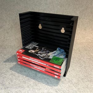 Op de afbeelding: Een zwarte, wandgemonteerde opbergrek voor videospelcartridges. Het rek bevat vier Nintendo Switch-spelcartridges, waaronder "Bravely Default II", "King's Bounty II", "Funky  Busters: Mastermind" en "Ancestors Legacy".