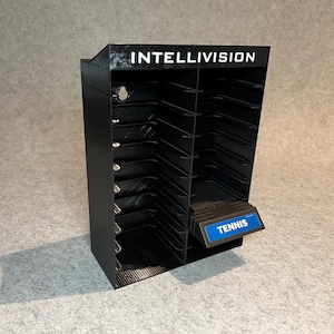 May include: A black plastic storage case for Intellivision video game cartridges. The case has 24 slots for cartridges and a pull-out drawer labeled "Tennis".