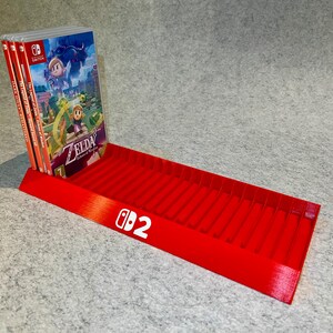 Puede incluir: Un soporte rojo brillante para cartuchos de juegos de Nintendo Switch. El soporte tiene un diseño inclinado con ranuras para cartuchos de juegos. El logotipo de Nintendo Switch y el número 2 están impresos en blanco. Se muestran varias carcasas de juegos.
