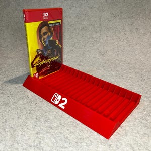 以下が含まれることがあります： 赤いプラスチック製のゲームカートリッジホルダー。Switchのロゴとゲームケースが付いています。ゲームケースには女性の肖像画と「Cyberpunk 2077」のタイトルが描かれています。ホルダーにはゲームカートリッジ用の複数のスロットがあります。