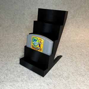 Op de afbeelding: Zwarte plastic standaard met vier niveaus voor het bewaren van Nintendo 64-spelcartridges. Een grijze cartridge met een geel label bevindt zich op het bovenste niveau. Het label luidt "Mario Tennis 64".