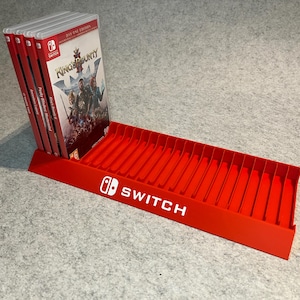 Op de afbeelding: Een rode plastic spelcassettehouder met het Nintendo Switch-logo in wit. Er worden verschillende gamecases weergegeven, met de speltitel "King's Bounty II" zichtbaar. De houder heeft meerdere sleuven voor gamecassettes.