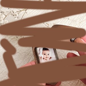 May include: A small silver picture frame with a photo of a baby inside. The baby is wearing a white onesie and has dark hair. The frame is sitting on a white surface.