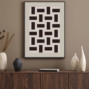 以下が含まれることがあります： 暗い茶色とベージュの長方形のブロックが交互に配置された幾何学模様の額入りアートプリント。 アートワークは、装飾的な花瓶と小さな本を備えた木製のキャビネットの上に展示されています。