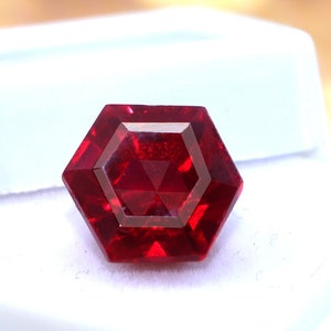 May include: A deep red gemstone with a hexagonal facet cut, displayed in a white box.