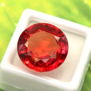 May include: A red gemstone with facets, set in a white square box. The box is on a green background.