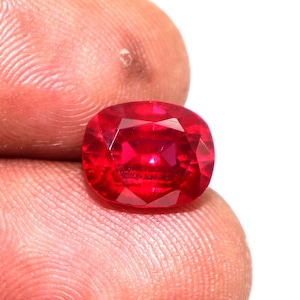 Puede incluir: Una piedra preciosa de color rojo intenso con talla cojín. La piedra preciosa se sostiene entre dos dedos.