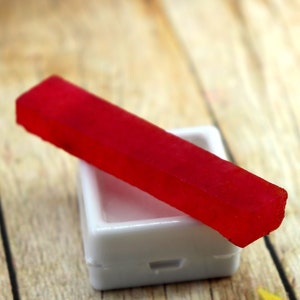 May include: A red watercolor paint stick in a white plastic container. The container has a small indentation on the top.