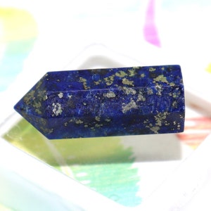 May include: A deep blue lapis lazuli crystal point with gold pyrite inclusions. The crystal is sitting on a white display stand.