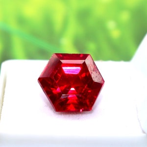 Könnte beinhalten: Ein tief roter Edelstein mit sechseckiger Form, in einer weißen Schachtel präsentiert. Der Edelstein ist facettiert und hat einen hohen Glanz.