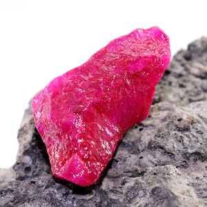 May include: A large, rough, pink ruby gemstone on a gray, textured rock.
