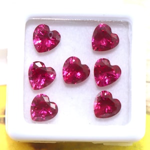 Puede incluir: Una caja blanca con seis piedras preciosas de rubí en forma de corazón. Las piedras preciosas son de color rosa intenso y están dispuestas en dos filas de tres.