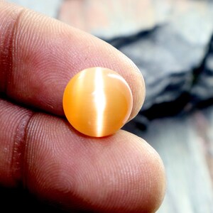 May include: A round, orange gemstone with a white, iridescent sheen. The gemstone is smooth and polished.