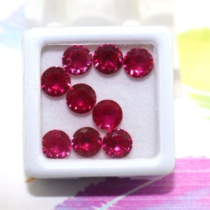 May include: A collection of ten round, red gemstones in a white box. The gemstones are arranged in a circular pattern.