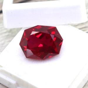 Pode incluir: Uma gema vermelha escura com muitas facetas é exibida em uma caixa quadrada branca. A caixa está sobre uma superfície de madeira.