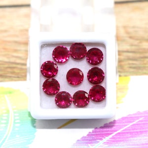 Könnte beinhalten: Nahaufnahme einer weißen Schachtel mit zehn runden, roten Edelsteinen. Die Edelsteine sind in einem Kreis angeordnet.