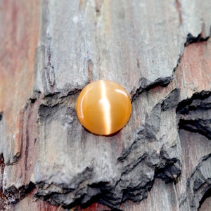 May include: A round, orange gemstone with a cat's eye effect. The gemstone is set against a rough, brown, textured background.