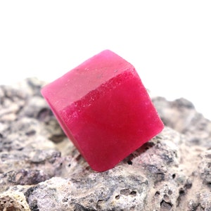 Może przedstawiać: Głęboko czerwony, fasetowany, sześcienny kryształ leży na szorstkiej, szarej, teksturowanej skale.