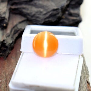May include: A round, orange gemstone with a bright, white, cat's eye effect. The gemstone is displayed in a white box.
