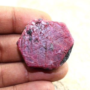 Puede incluir: Una piedra preciosa de rubí rojo intenso con forma hexagonal y una superficie rugosa y facetada. El rubí tiene un aspecto natural y sin pulir.