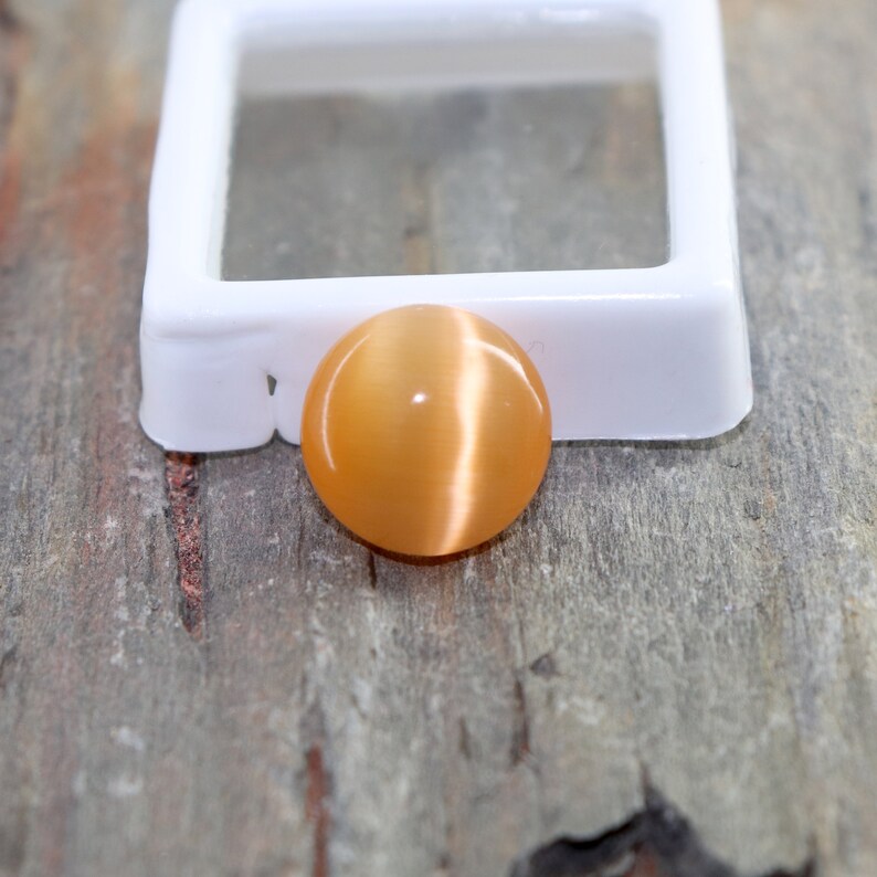 May include: A round, orange gemstone with a chatoyant effect. The gemstone is set in a white, square display box.