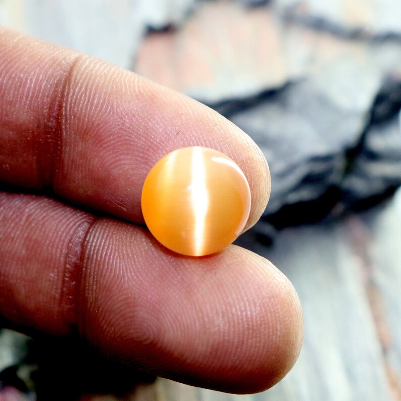 May include: A round, orange gemstone with a white, iridescent sheen. The gemstone is smooth and polished.