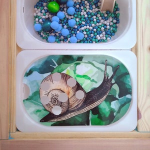 Może przedstawiać: Biała taca z ilustracją ślimaka na tle zielonych liści. Druga taca zawiera łopatkę i mieszankę białych, niebieskich i zielonych koralików.