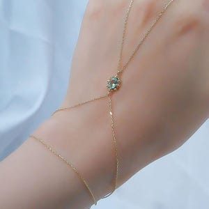 Puede incluir: Delicada cadena de mano dorada con una piedra preciosa central redonda y verde claro. La cadena se conecta a un anillo y se envuelve alrededor de la mano, creando una elegante y minimalista pieza de joyería. El fondo es una tela blanca suave.