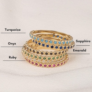 May include: Five gold rings with different coloured gemstones. The rings are stacked on top of each other. The gemstones are turquoise, sapphire, onyx, emerald, and ruby.