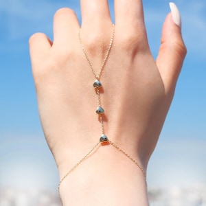 May include: A delicate gold hand chain featuring three heart-shaped charms. The chain extends from the wrist to the middle finger, creating a stylish and elegant accessory. The background is a bright blue sky.