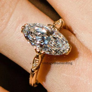 May include: A gold ring with a large, oval-shaped diamond. The diamond is clear and sparkling, set in a gold band with decorative details. The ring is on a finger.