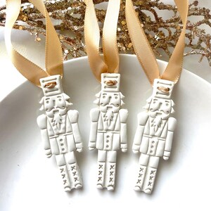May include: Three white ceramic nutcracker ornaments with gold accents and a gold ribbon. The ornaments are hanging from the ribbon.