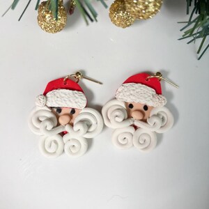 Könnte beinhalten: Ein Paar festliche Santa Claus-Ohrringe. Jeder Ohrring hat einen roten Hut mit weißem Rand, ein rosiges Gesicht und einen weißen, lockigen Bart und Schnurrbart. Die Ohrringe sind aus Polymer Clay gefertigt und haben goldfarbene Ohrhaken. Perfekt für Weihnachten.