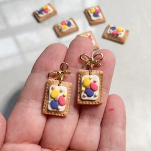 May include: Handmade gold-toned bow earrings with miniature biscuit charms. The biscuit charms have a brown border, white frosting, and colorful candy toppings. Additional biscuit charms are scattered in the background.