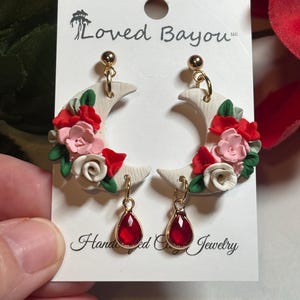 May include: Handmade crescent moon earrings with red, pink, and white floral accents. Each earring features a gold-colored teardrop-shaped red gem. The earrings are attached to a white card with the text "Loved Bayou" and "Handmade Clay Jewelry."