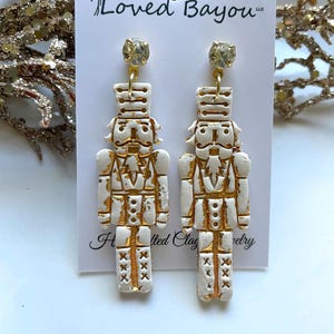 May include: White porcelain nutcracker earrings with gold accents. The earrings are dangle style and feature a rhinestone at the top.