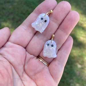 May include: A pair of ghost-shaped earrings featuring a translucent, iridescent finish with embedded glitter particles. Each ghost has two small, circular black eyes and is attached to a gold-toned metal stud post with a small jump ring connector.