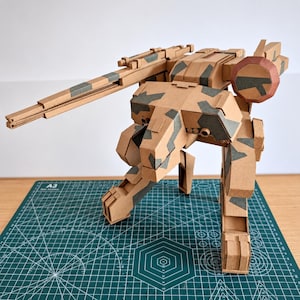 Puede incluir: Un modelo de cartón de un robot bípedo con un patrón de camuflaje en color tostado y gris oscuro. El robot presenta un brazo de cañón largo y prominente, un componente lateral circular y estructuras de patas articuladas de cartón marrón.
