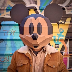 Peut inclure: Une personne portant un grand masque géométrique de Mickey Mouse en carton. Le masque est marron, noir et beige. La personne porte une veste en velours côtelé marron avec un col en peau de mouton blanc et une chemise boutonnée beige. Fond de graffiti.