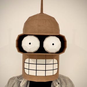 May include: A brown cardboard Bender mask from the animated series Futurama. The mask features large white eyes with black pupils, a wide grin with white teeth, and a tall antenna. The mask is worn over a person's head.