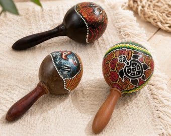 May include: Three wooden maracas with rounded heads and smooth handles. One features a black finish with red and orange accents, another displays a blue and brown design, and the third showcases a green and red pattern with a white sea turtle.
