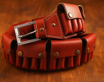 May include: A red leather belt with multiple cartridge pouches. The belt features a silver buckle and brass-colored rivets. The pouches are designed to hold ammunition. The belt is displayed on a wooden surface.
