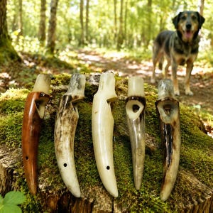 May include: Five handcrafted bone whistles of varying colours and textures are displayed on a moss-covered tree stump. The whistles have a smooth, polished finish and a hole near the base. A dog stands in the background.