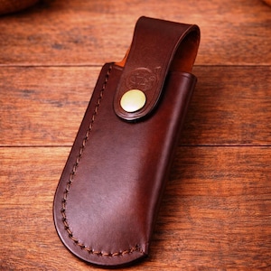 May include: A brown leather knife sheath with a curved bottom and a flap secured by a brass-colored button. The sheath is stitched with brown thread and rests on a wooden surface.