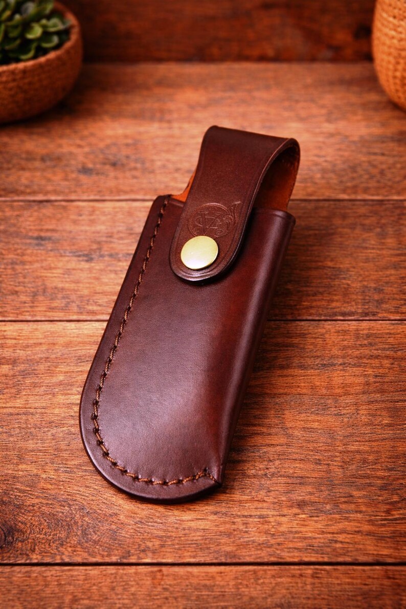 May include: A brown leather knife sheath with a curved bottom and a flap secured by a brass-colored button. The sheath is stitched with brown thread and rests on a wooden surface.