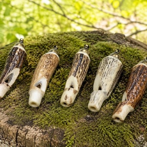 May include: Five whistles crafted from natural antler, displaying varied brown and cream patterns. They are arranged on a moss-covered tree stump. The background is a blurred green, suggesting a natural setting.