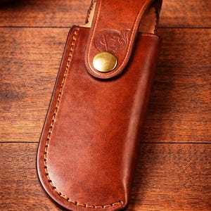 May include: A brown leather knife sheath with a flap closure and a brass button. The sheath is stitched with a contrasting thread and features a decorative emblem on the flap. The leather has a rich, warm tone.